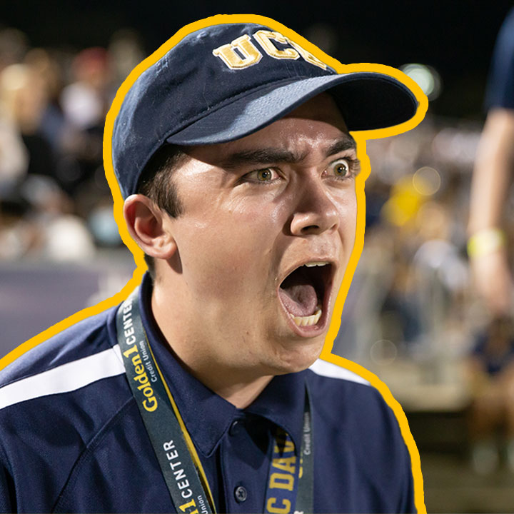 A spirited UC Davis student, clad in university colors and a baseball cap, shouts with fervor, surrounded by the bustling energy of fellow fans at the Homecoming football game.