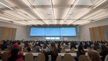 large lecture room with three large tv screens and rows of college students in chairs