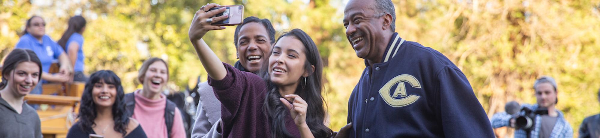 Student laughing while posing with selfie with Chancellor May and LeShelle May on the quad