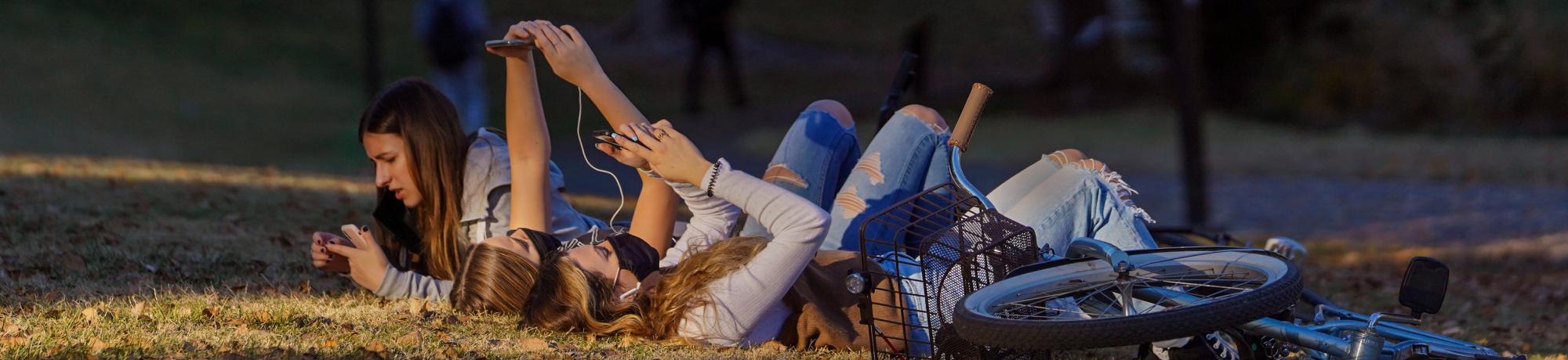 A group of students wearing face masks and lying on grass watch videos on their smartphones near Lake Spafford in the UC Davis Arboretum.